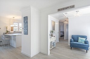 A modern kitchen with a bar area, a dining area with a blue armchair, and a hallway leading to other rooms. - Sunny Days: Bracklesham Bay Beach Bungalow – Sleeps 7, Parking & Dog Friendly (Bracklesham Bay)