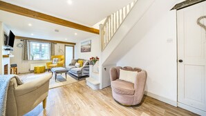 A spacious living room with a staircase, wooden beams, and a large window. - West Wittering Rosemary Cottage – Sleeps 5, Nr Beach, Parking & Family Friendly (West Wittering)