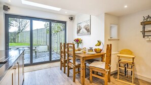 A modern kitchen with a dining area, large sliding glass doors to a garden, and a wooden table set for breakfast. - West Wittering Rosemary Cottage – Sleeps 5, Nr Beach, Parking & Family Friendly (West Wittering)