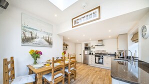A modern kitchen with a central island, wooden dining table with chairs, and a large painting on the wall. - West Wittering Rosemary Cottage – Sleeps 5, Nr Beach, Parking & Family Friendly (West Wittering)