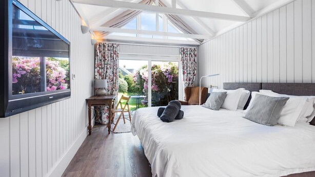 Ein Schlafzimmer mit einem großen Bett, einem an der Wand montierten Fernseher und Blick auf einen Garten durch das Fenster.