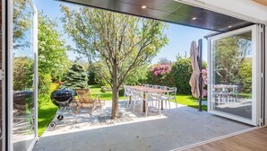 Eine Terrasse mit Grill, Tisch und Stühlen sowie eine Glastür, die zu einem Garten mit Bäumen und Blumen führt.