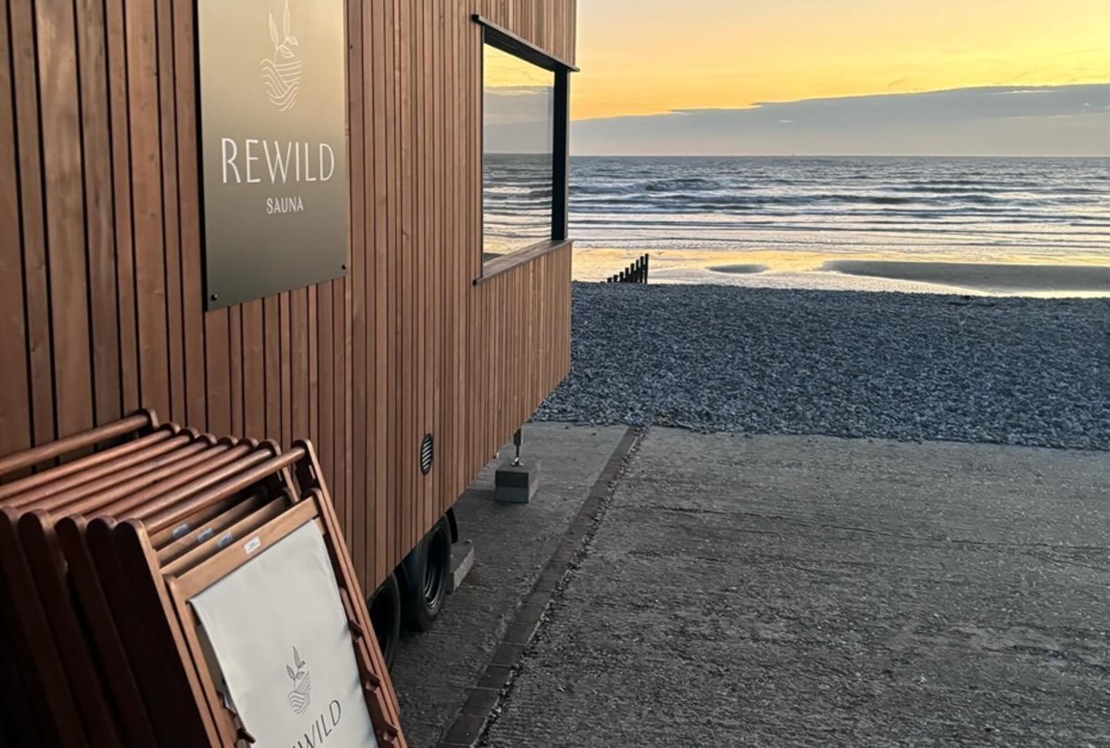A wooden building with a sign reading 'REWILD SAUNA' and a view of the ocean.