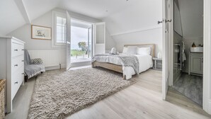 A bedroom with a large window, a bed, a dresser, and a view of the outdoors. - Seacote: Family Seafront Home in Selsey – Sleeps 7, Hot Tub & Sea Views (Selsey)