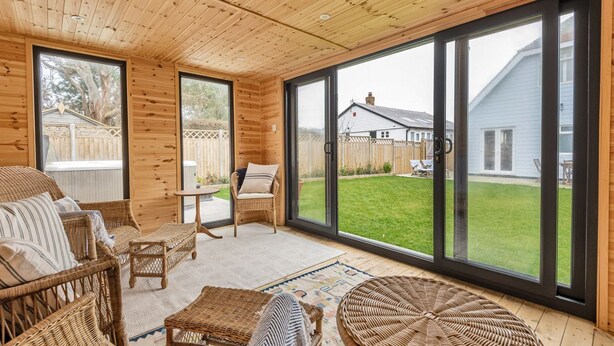 A cozy room with wooden walls and large windows, wicker furniture, and a view of a house and yard. - Seacote: Family Seafront Home in Selsey – Sleeps 7, Hot Tub & Sea Views (Selsey)