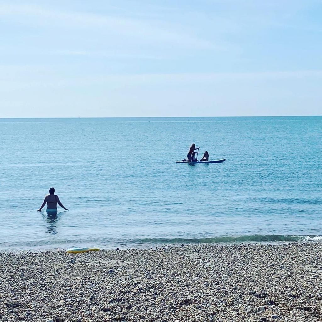 Une plage de galets avec une eau calme, une personne debout dans l’eau et deux autres personnes qui pratiquent le paddleboard au loin.