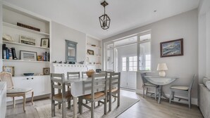 A dining area with a round table, chairs, and a white cabinet with shelves. - Seacote: Family Seafront Home in Selsey – Sleeps 7, Hot Tub & Sea Views (Selsey)