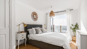Un dormitorio con una cama grande, una mesita de noche, una lámpara, un reloj de pared y vistas al mar a través de una ventana.