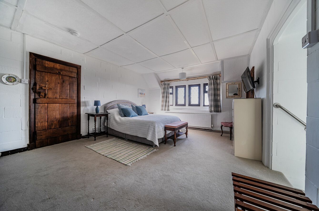 Un dormitorio con una puerta grande de madera, una cama con cubrecama blanco, una mesita de noche con lámpara, un radiador y una ventana con cortinas.