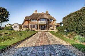 A large, two-story house with a thatched roof, a paved driveway, and well-maintained gardens. - Beachfront Thatched Cottage in West Wittering – Sleeps 8, Garden, Parking, Pets (West Wittering)