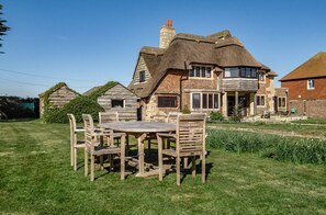A large house with a thatched roof, a stone chimney, and a wooden dining table with chairs in the front yard. - Beachfront Thatched Cottage in West Wittering – Sleeps 8, Garden, Parking, Pets (West Wittering)
