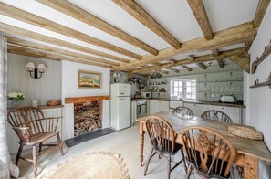 A rustic kitchen with wooden beams, a dining table, chairs, and a fireplace. - Charming West Wittering Cottage – Cosy Village Retreat, Pet Friendly (West Wittering)