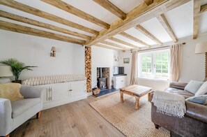 A cozy living room with exposed wooden beams, a fireplace, a sofa, a coffee table, and a view of greenery outside. - Charming West Wittering Cottage – Cosy Village Retreat, Pet Friendly (West Wittering)