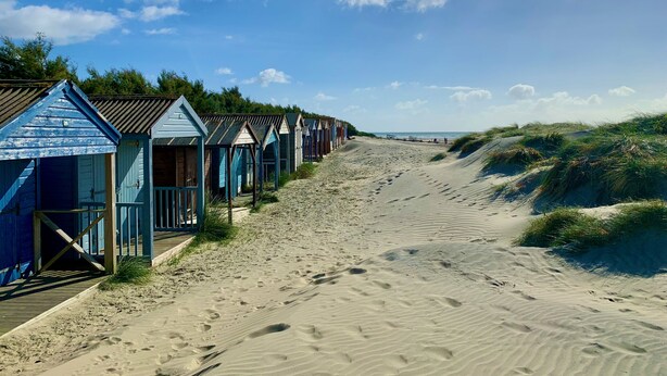A row of beach huts with a sandy path leading to the sea. - Stylish West Wittering Retreat Near Beach – Thessaly, Sleeps 8, Pet Friendly (West Wittering)