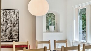 A dining area with a wooden table, chairs, a hanging light fixture, a framed black and white photograph, and a window with a view of greenery. - Stylish West Wittering Retreat Near Beach – Thessaly, Sleeps 8, Pet Friendly (West Wittering)