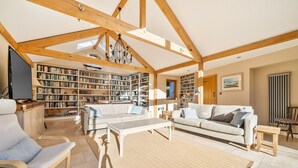 A spacious living room with a high ceiling, exposed wooden beams, a large bookshelf, a sofa, and a coffee table. - West Wittering Family Beach Escape – Sleeps 8, Garden & Dog Friendly (West Wittering)
