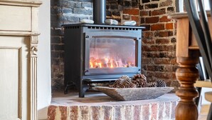 A wood-burning stove with a brick wall and a wooden bowl of pinecones. - West Wittering Family Beach Escape – Sleeps 8, Garden & Dog Friendly (West Wittering)
