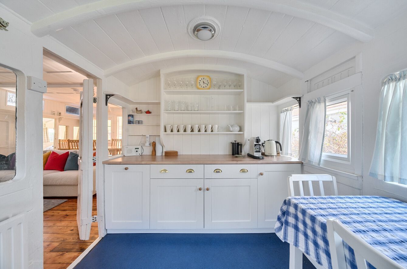 Een compacte keuken met witte kastjes, een blauw-wit geruit tafelkleed en een raam met dunne gordijnen.