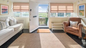 A cozy living room with a white sofa, a brown leather armchair, and a coffee table. There are two windows with patterned curtains, a door leading outside, and a view of a garden. - Unique Railway Carriage Home on Selsey Seafront – Sleeps 7, 5 Bedrooms (Selsey)