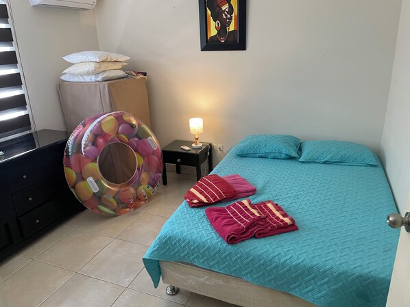 A bedroom with a bed, pillows, a lamp, a trash bin, and a colorful inflatable ball.