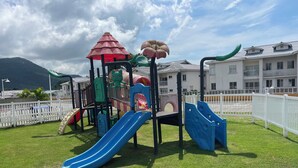 Ein Spielplatz mit Rutschen und Klettergerüsten, umgeben von einem weißen Zaun und Gebäuden im Hintergrund.