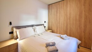 Een keurig opgemaakt bed met witte lakens en een opgevouwen handdoek, een nachtkastje van hout met een lamp, en een houten garderobe tegen de muur.