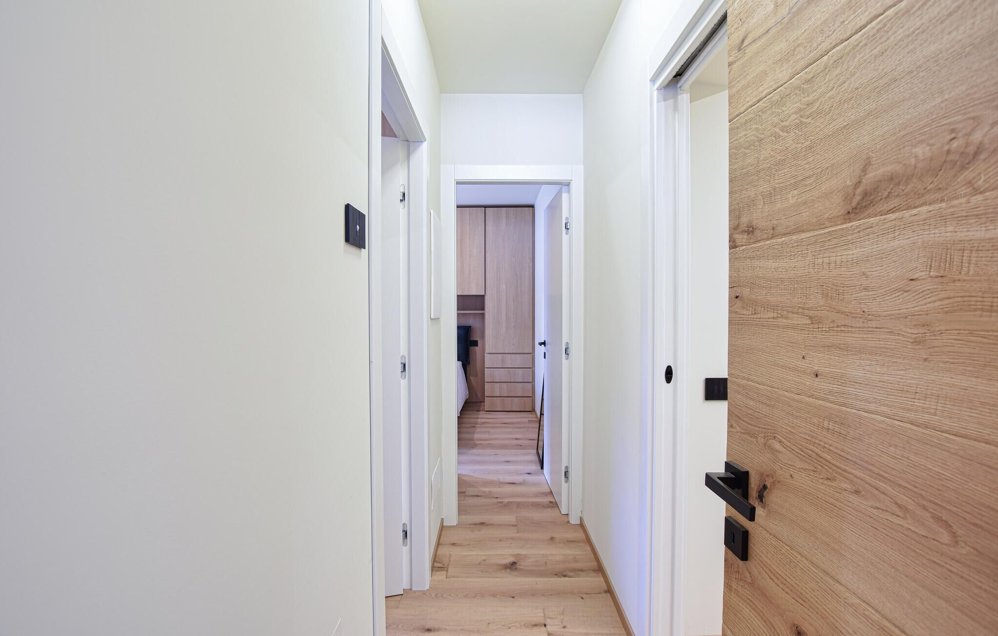 Un pasillo estrecho con suelo de madera, paredes blancas y una puerta de madera a la derecha.