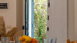 Un comedor con una mesa lista para dos personas, un frutero y vistas a un jardín a través de una puerta abierta.