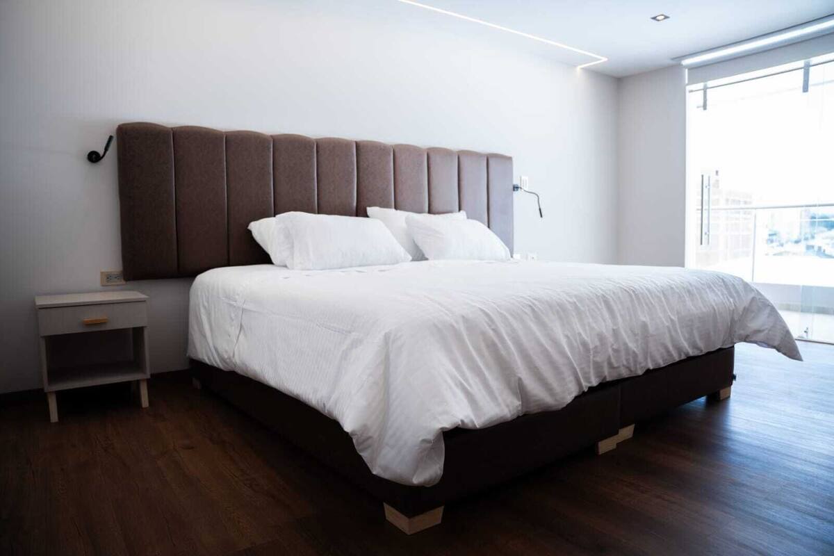 Un dormitorio moderno con una cama grande, una mesita de noche y vistas de la ciudad a través de un ventanal amplio.
