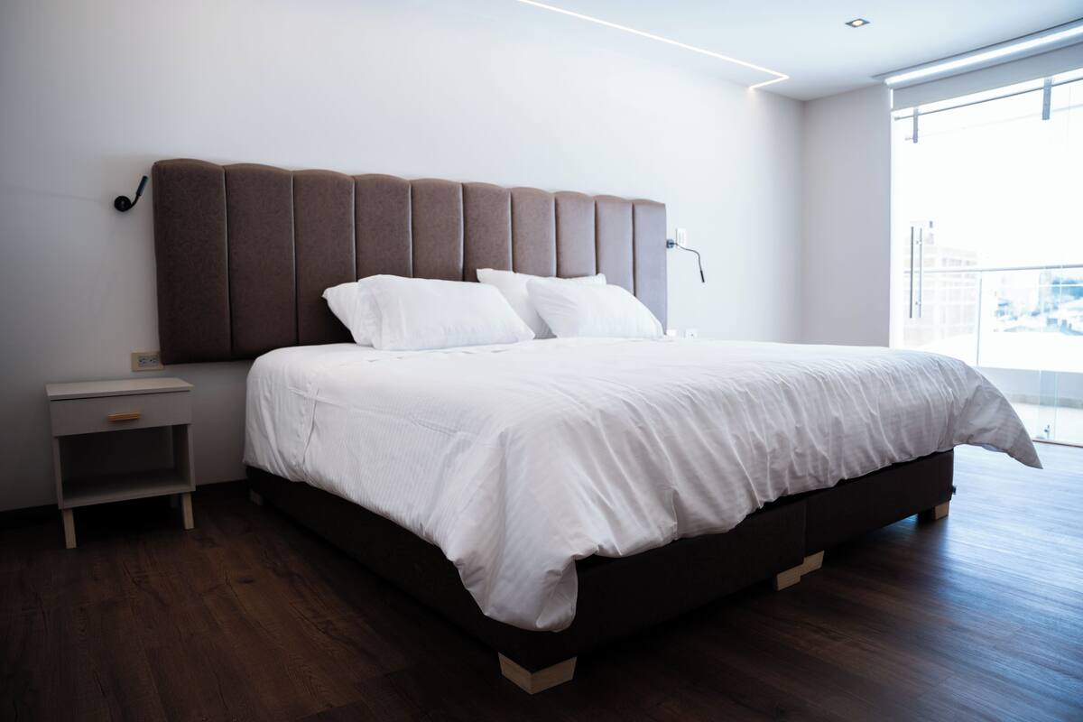 Un dormitorio moderno con una cama grande, una mesita de noche y vistas de la ciudad a través de un ventanal amplio.