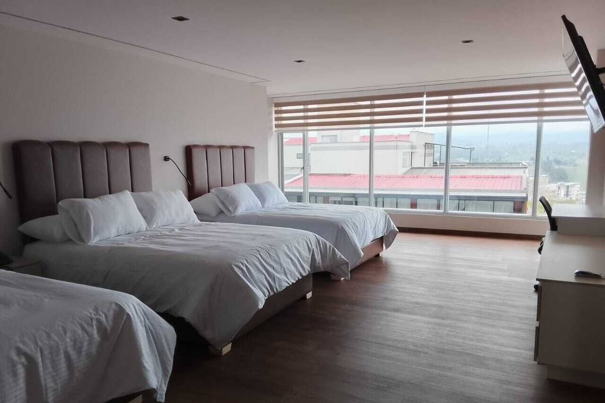 Habitación de hotel con tres camas, un ventanal con persianas y suelo de madera.