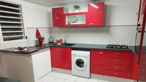 Een kleine keuken met rode kastjes, een ingebouwde wasmachine en een raam met zonwering.