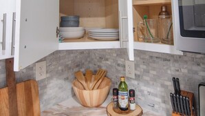 Uma cozinha com armários brancos, micro-ondas e bancada com tábua de corte de madeira, porta-facas e um frasco de azeite.