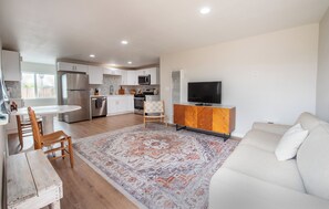A modern kitchen with built-in appliances, a dining area with a wooden table and chairs, a living room with a sofa and a flat-screen TV, and a patterned rug on the floor. - Grover Beach Getaway near beach, downtown & dunes! (Grover Beach)