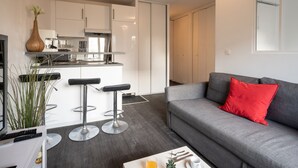 Ein zeitgemäßer Wohnbereich mit grauem Sofa, rotem Kissen und einer Küchenzeile mit weißen Schränken sowie einem Barhocker.