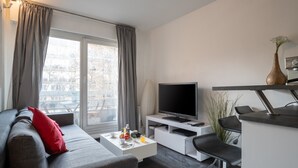 Ein modernes Wohnzimmer mit einem grauen Sofa, einem Couchtisch mit Früchten und einer Fernbedienung, einem Fernseher auf einem weißen Schrank sowie einer Küchenzeile mit einer Theke und Stühlen.