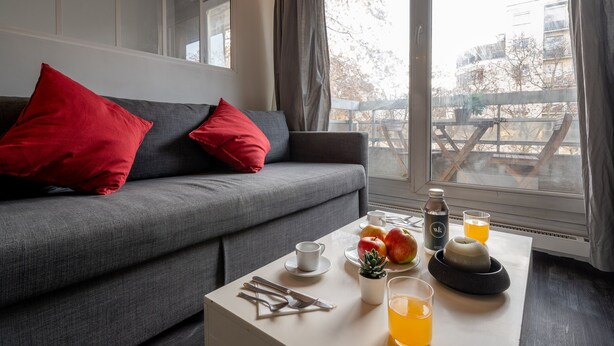 Ein modernes Wohnzimmer mit einem grauen Sofa, roten Kissen, einem Esstisch mit Frühstücksgebäck und Blick auf einen Balkon mit Gartenmöbeln.