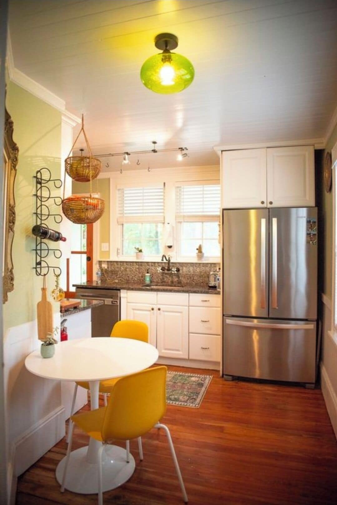 Una cocina compacta con armarios blancos, un refrigerador de acero inoxidable, una mesa de comedor con dos sillas y una lámpara colgante.
