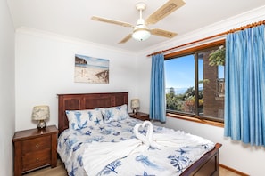 A bedroom with a bed, wooden headboard, nightstands, a ceiling fan, a window with curtains, and a beach painting on the wall. - Wallawa Water Views Family Retreat in Nelson Bay (Nelson Bay)