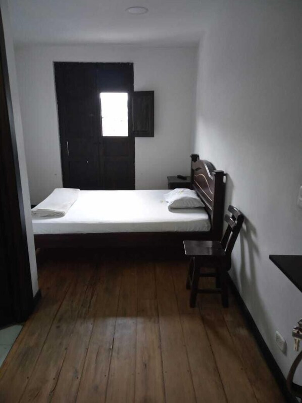 Une petite chambre avec un lit simple, un plancher en bois, un bureau et une chaise.