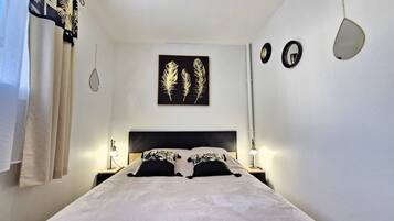 Une chambre à coucher comprenant un lit, deux tables de chevet, un miroir fixé au mur et une œuvre d’art encadrée représentant des plumes.