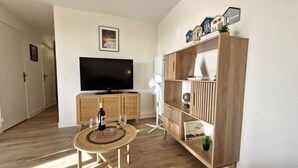 A living room with a flat-screen TV, a wooden cabinet, a small round table with a bottle and glasses, and a standing fan. - Royan – Fully Renovated  et  Fully Equipped Apartment – Perfect for Holidays! (ROYAN)
