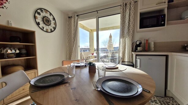 A dining area with a wooden table set for two, a clock on the wall, and a view of the outside through a large window. - Royan – Fully Renovated  et  Fully Equipped Apartment – Perfect for Holidays! (ROYAN)