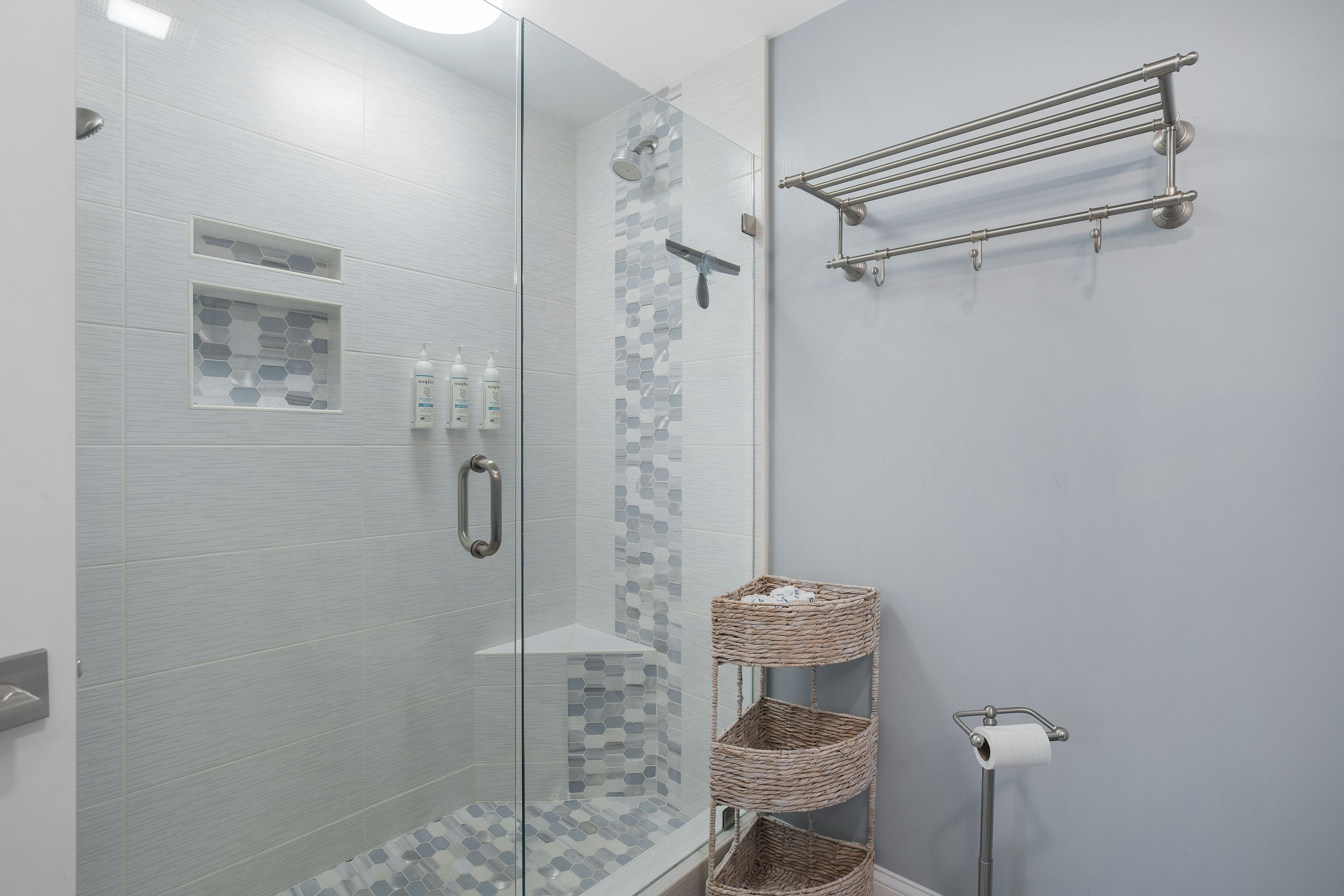 A modern bathroom with a glass-enclosed shower, a white tiled wall, a small shelf with toiletries, and a wicker basket.