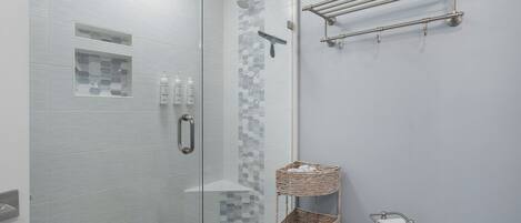 A modern bathroom with a glass-enclosed shower, a white tiled wall, a small shelf with toiletries, and a wicker basket.