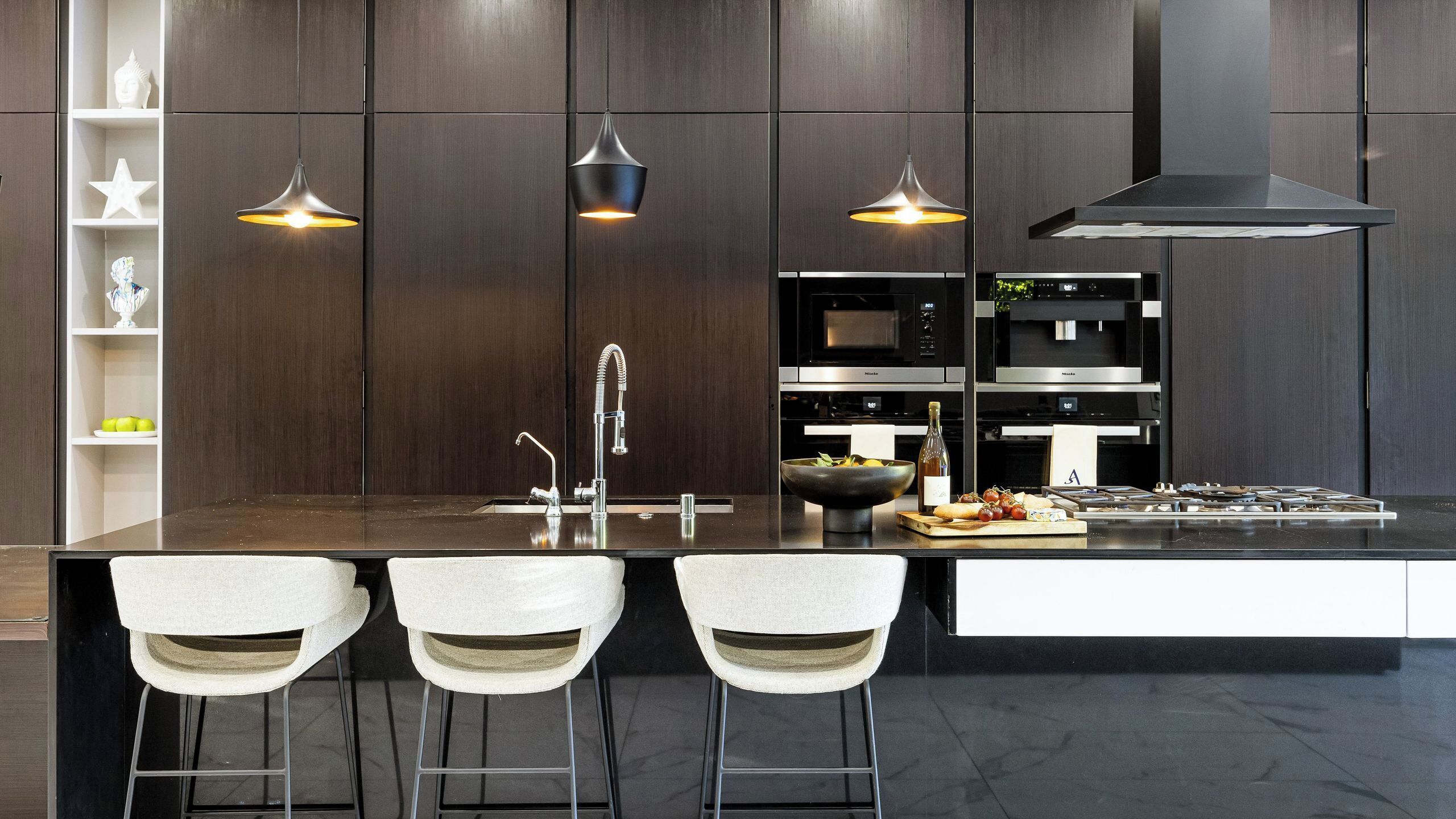 A modern kitchen with a central island, bar stools, pendant lighting, stainless steel appliances, and a dark wood backsplash.