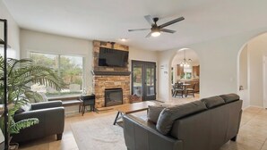 A spacious living room with a stone fireplace, a large grey sofa, a flat-screen TV mounted above the fireplace, and a ceiling fan.