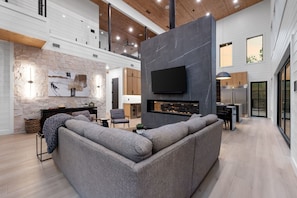 A modern living room with a grey sectional sofa, a fireplace, and a flat-screen TV mounted on a dark wall. - Luxury Cabin w/ Golf Sim & Pickleball Court (Broken Bow)