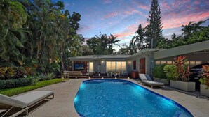 A swimming pool with lounge chairs and a patio area, surrounded by tropical plants and trees. - High-End House with Heated Pool, outdoor TV (Miami Shores)