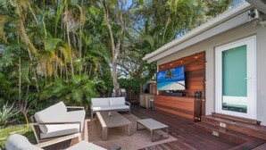 An outdoor living area with a wooden deck, white furniture, and a TV mounted on the wall. - High-End House with Heated Pool, outdoor TV (Miami Shores)
